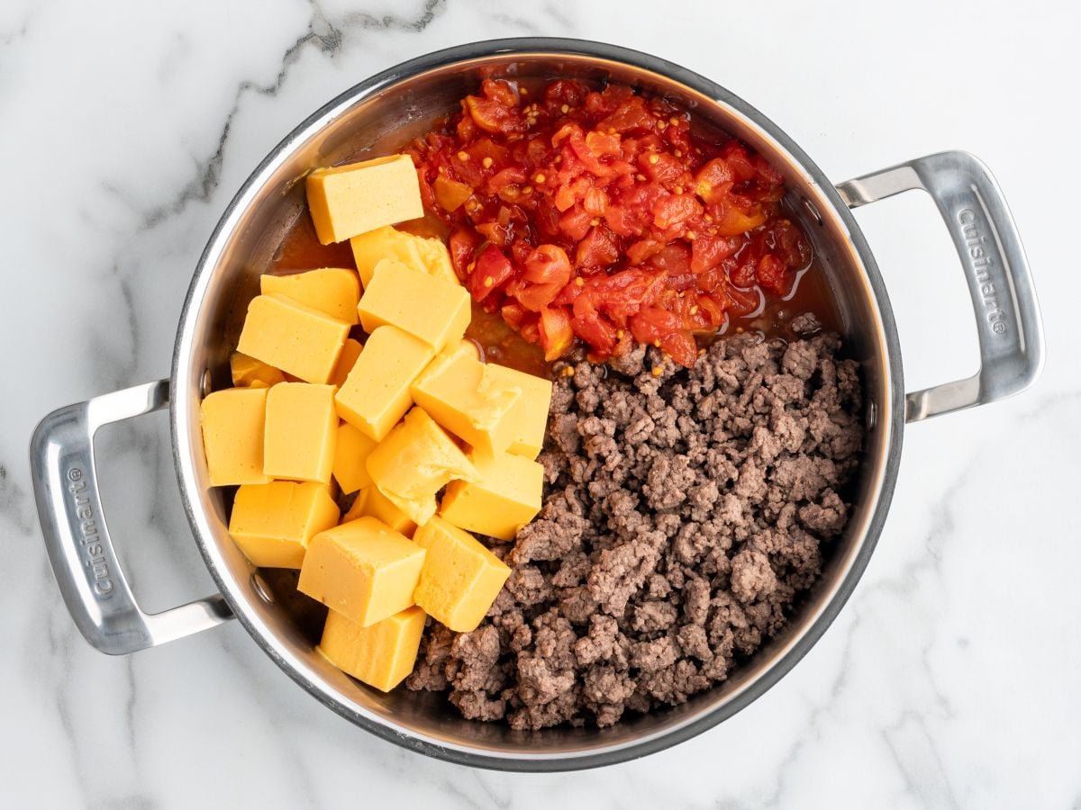A stainless steel pan on a marble surface holds cubed yellow cheese, cooked ground beef, and Rotel tomatoes with green chilies, arranged separately before mixing into a classic Rotel Dip.