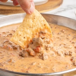 A hand dips a tortilla chip into a skillet filled with creamy Rotel dip containing ground beef and diced tomatoes.