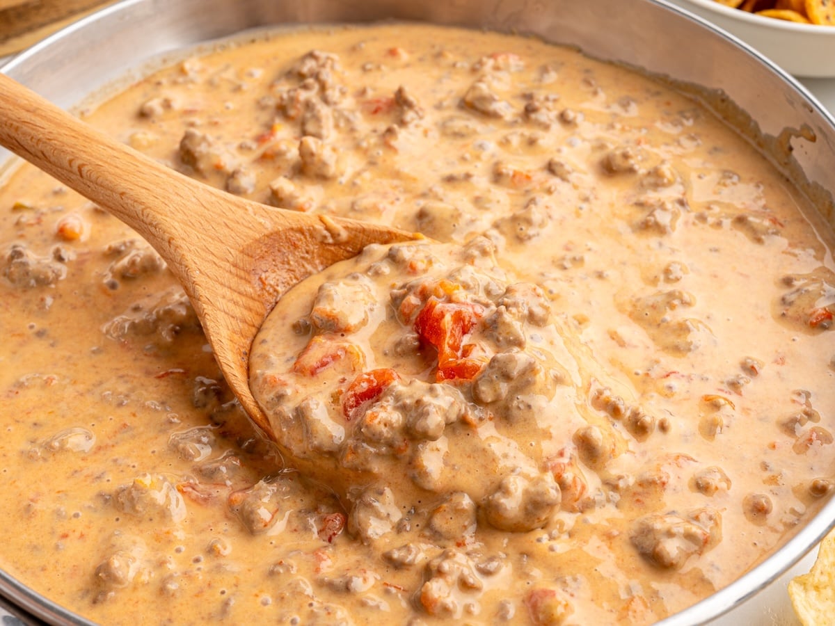 A wooden spoon stirs a creamy, chunky Rotel dip with ground beef and diced tomatoes in a pan.
