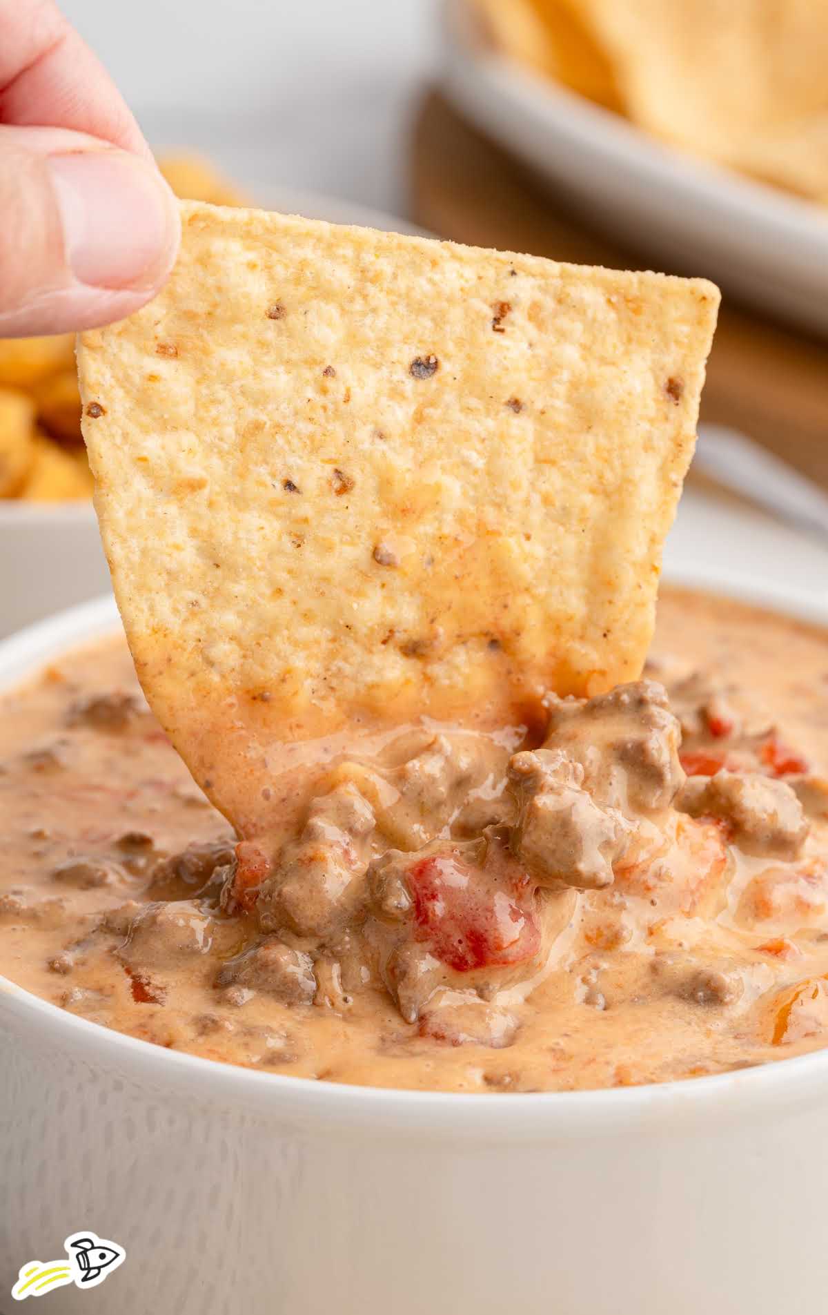 Hand dipping a tortilla chip into a bowl of creamy beef and tomato Rotel dip.
