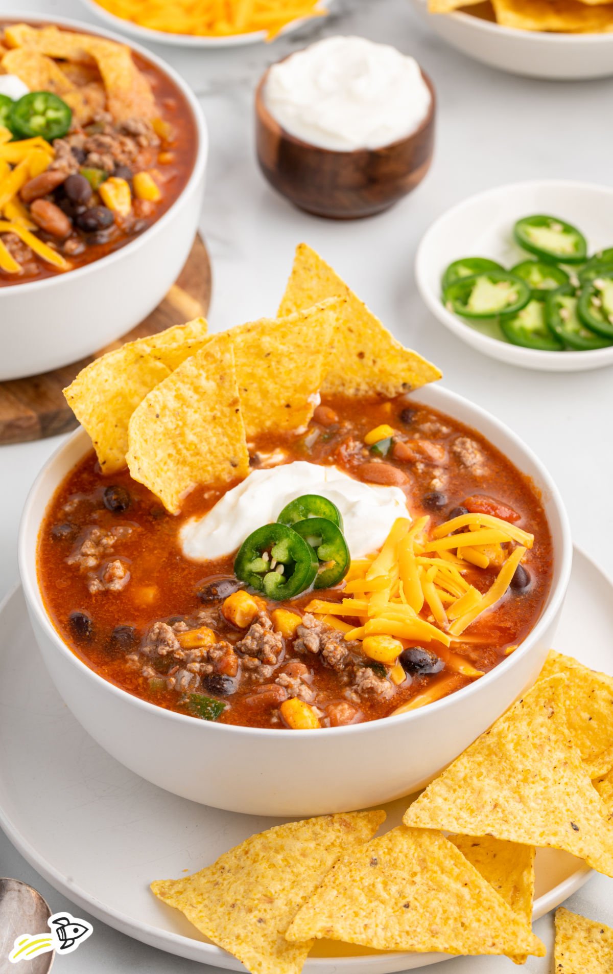 A vibrant bowl of taco soup garnished with shredded cheese, sour cream, jalapeño slices, and tortilla chips arranged along the rim. The bowl sits on a white plate with extra tortilla chips beside it, with small bowls of sliced jalapeños and sour cream in the background.