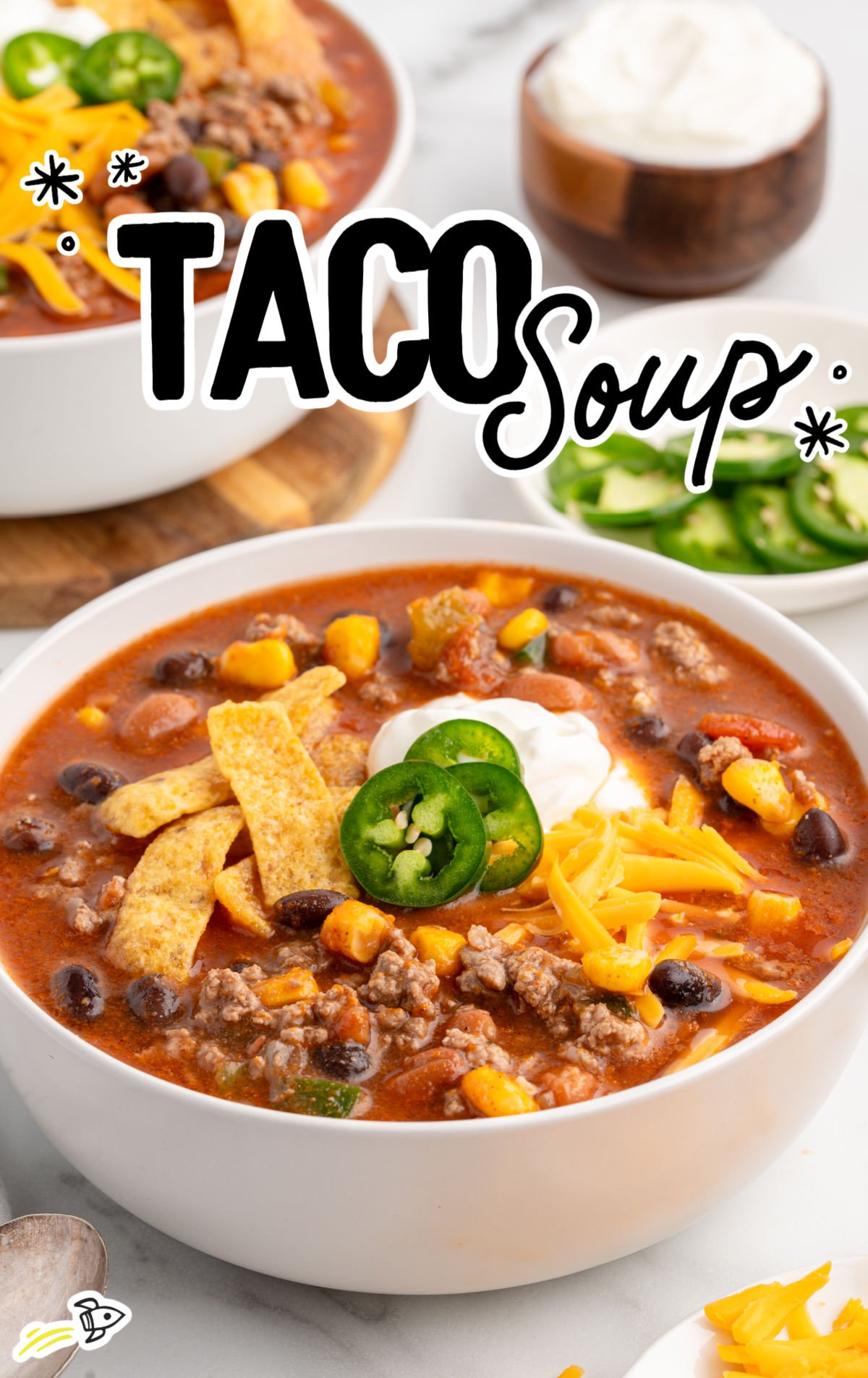A close-up bowl of taco soup topped with shredded cheddar cheese, sour cream, jalapeño slices, corn chips, and surrounded by sides of jalapeños and sour cream. The rich tomato broth is filled with ground beef, beans, corn, and visible seasonings, styled on a light backdrop.