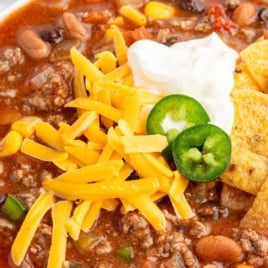 Vertical shot of taco soup with a topping of cheese, sour cream, jalapeños, and tortilla chips; text overlay reads “Easy & Filling Taco Soup – Just Dump the Cans In & Simmer!”