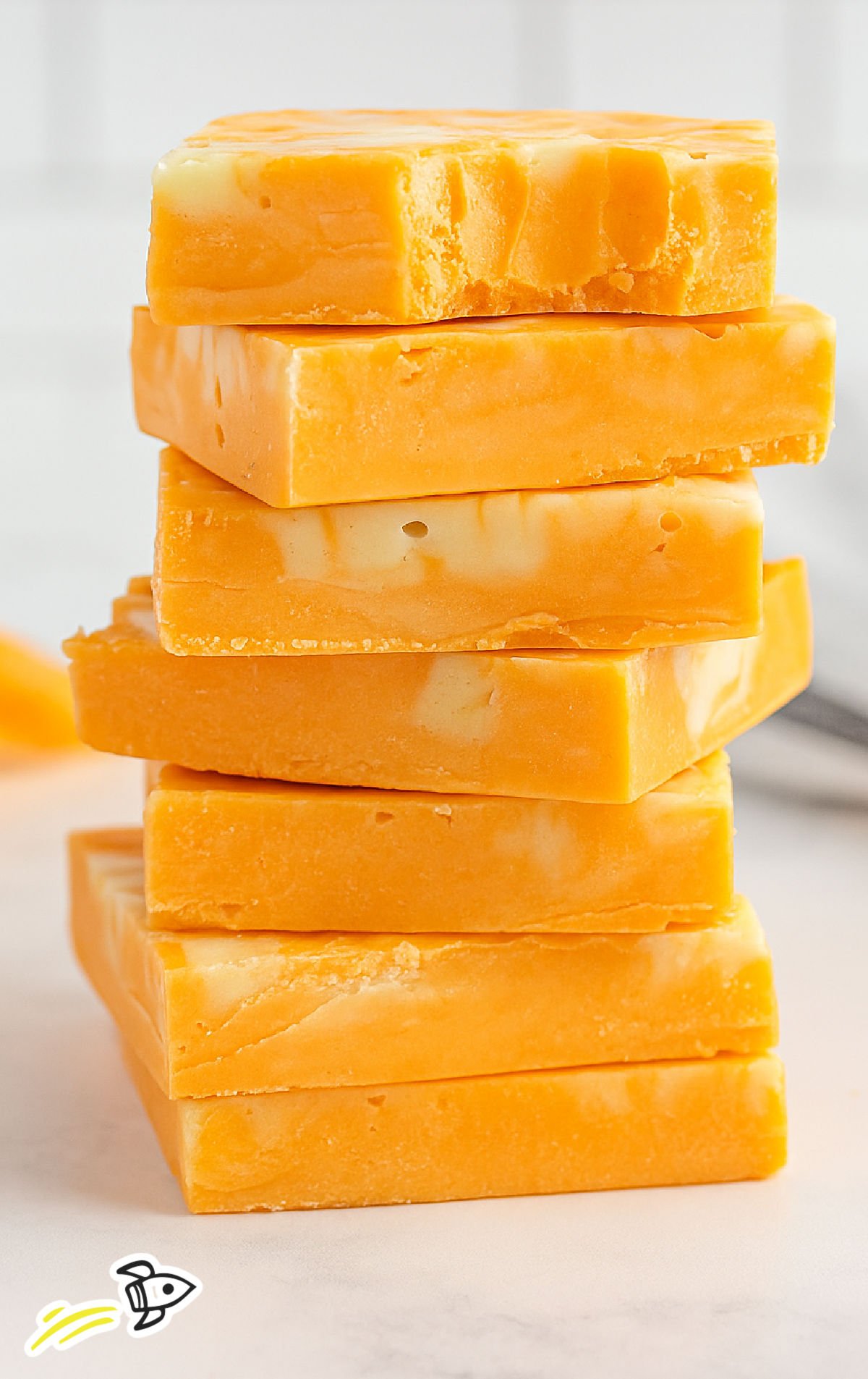 Stack of orange and white swirled creamsicle fudge squares on a light marble surface, with the top piece showing a small bite taken out to reveal its creamy texture. The background is softly blurred with warm, natural lighting