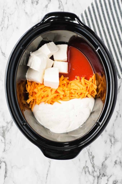 A Crockpot Buffalo Chicken Dip Recipe featuring blocks of cream cheese, shredded cheddar, ranch dressing, and buffalo sauce sits in a slow cooker on a marble countertop with a striped cloth nearby, all ingredients ready to be mixed.