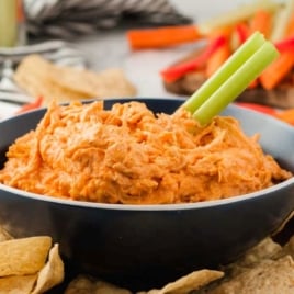 A black bowl filled with creamy orange Crockpot Buffalo Chicken Dip, garnished with a celery stick and surrounded by tortilla chips. Sliced vegetables are visible in the background.
