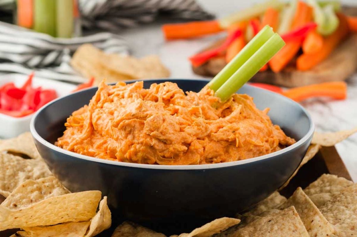 A black bowl filled with creamy orange Crockpot Buffalo Chicken Dip, garnished with a celery stick and surrounded by tortilla chips. Sliced vegetables are visible in the background.