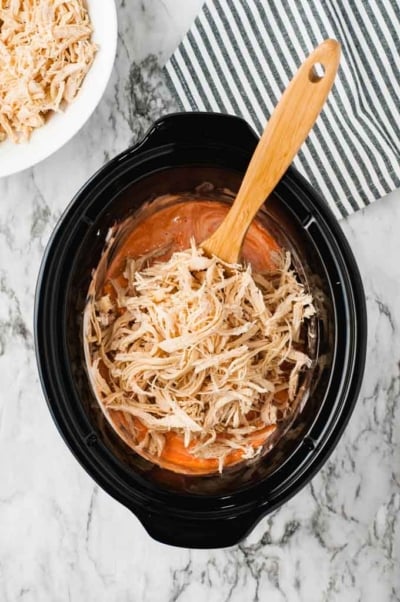 Overhead view of a slow cooker filled with shredded chicken and sauce for a Crockpot Buffalo Chicken Dip Recipe, being stirred with a wooden spoon. Nearby, a bowl of more shredded chicken and a striped napkin sit on the marble countertop.