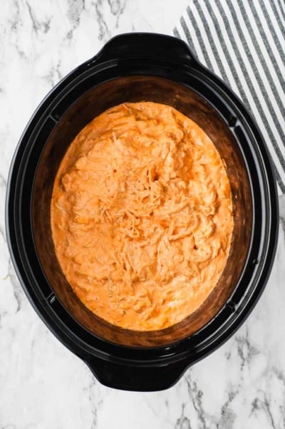 Crockpot Buffalo Chicken Dip in a slow cooker, featuring a creamy, orange mixture with shredded chicken, set on a marble countertop next to a black and white striped cloth.