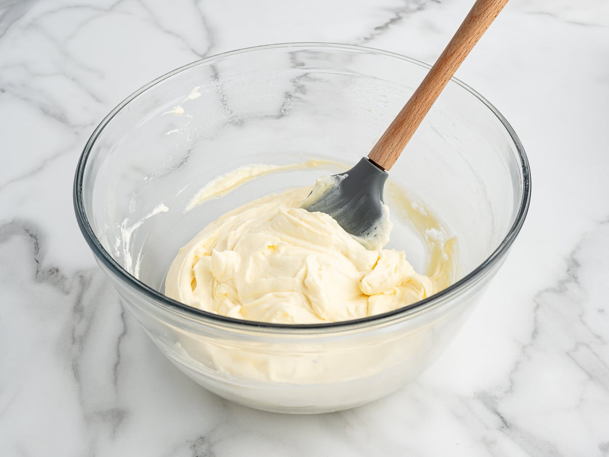 A glass bowl filled with creamy, whipped Banana Pudding Pie batter sits on a marble countertop, with a spatula resting inside the bowl.