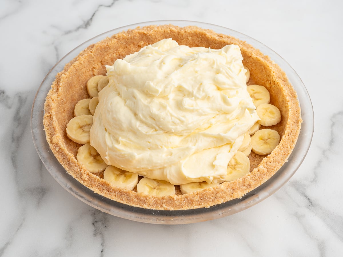 A Banana Pudding Pie with a graham cracker crust, layered with sliced bananas and topped with a large mound of creamy, pale yellow filling, sits on a marble surface.