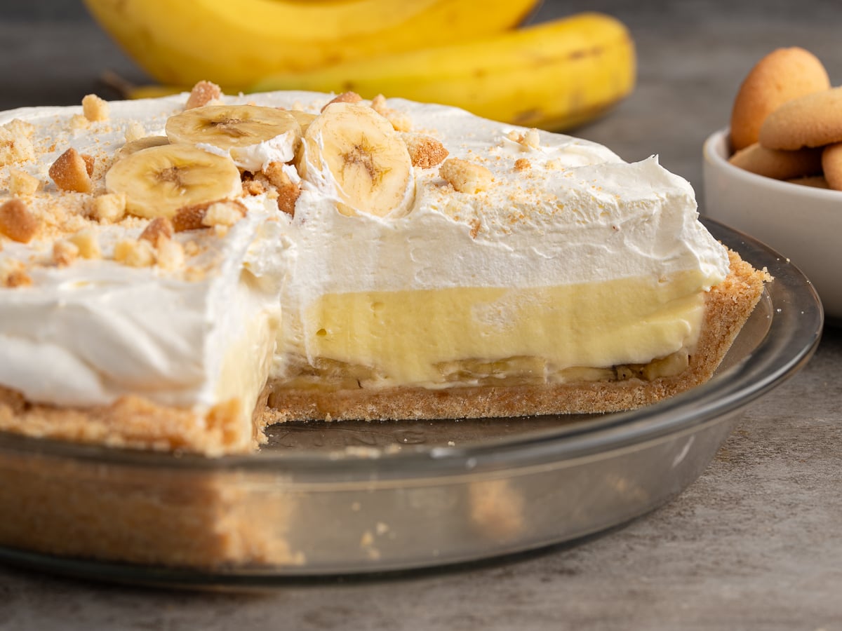 A Banana Pudding Pie with a graham cracker crust, topped with whipped cream, banana slices, and crumbled cookies. A slice has been removed, revealing creamy filling and bananas inside. Bananas and cookies sit in the background.