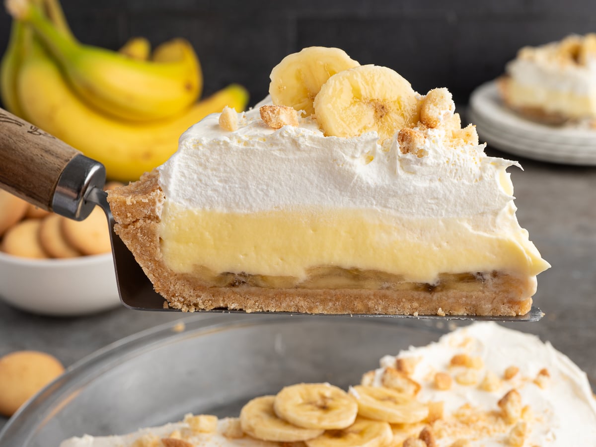 A close-up of a Banana Pudding Pie slice on a pie server, showing layers of crust, banana, creamy filling, and whipped topping, garnished with banana slices and crumbled cookies. Whole bananas and pie appear in the background.