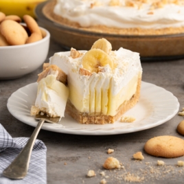 A slice of Banana Pudding Pie with whipped topping and banana slices sits on a white plate, with a forkful cut out. Crushed vanilla wafers and a bowl of whole wafers are nearby, while the whole pie rests in the background.