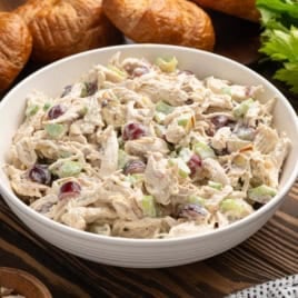 A bowl of creamy chicken salad with shredded chicken, celery, and red grapes sits on a wooden table, perfect for making a delicious Chicken Salad Sandwich. Croissants and fresh celery are in the background.
