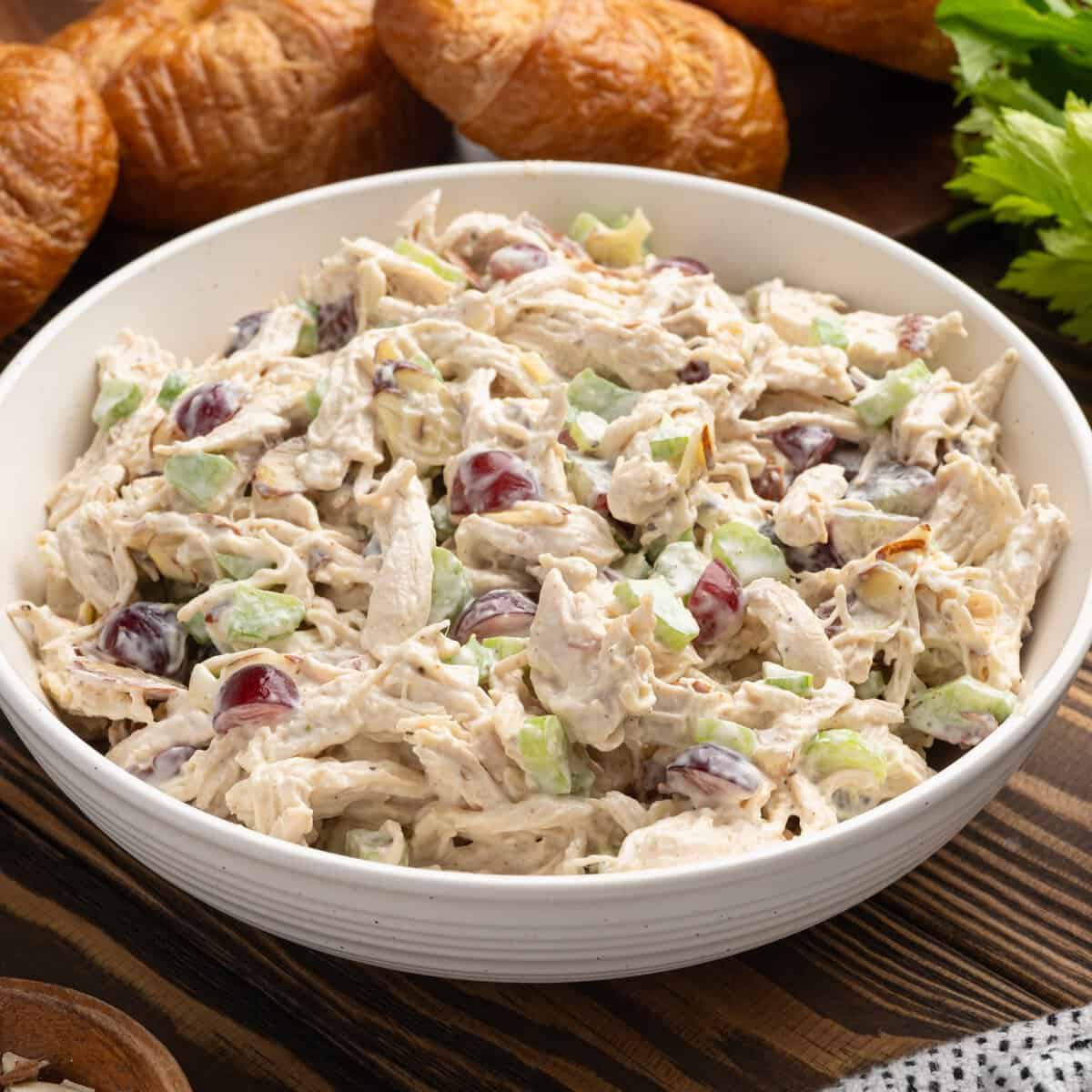 A bowl of creamy chicken salad with shredded chicken, celery, and red grapes sits on a wooden table, perfect for making a delicious Chicken Salad Sandwich. Croissants and fresh celery are in the background.