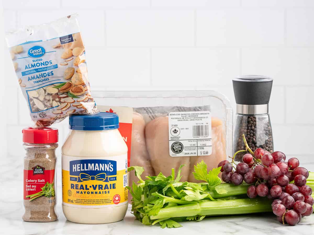 A selection of ingredients for a Chicken Salad Sandwich on a counter: sliced almonds, celery salt, mayonnaise, celery stalks, raw chicken breasts, black pepper grinder, and a bunch of red grapes.