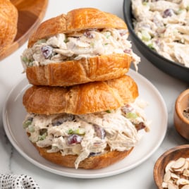 Two Chicken Salad Sandwich croissants filled with creamy chicken salad, grapes, celery, and almonds are stacked on a white plate, surrounded by a bowl of salad, grapes, sliced almonds, and pepper on a marble surface.