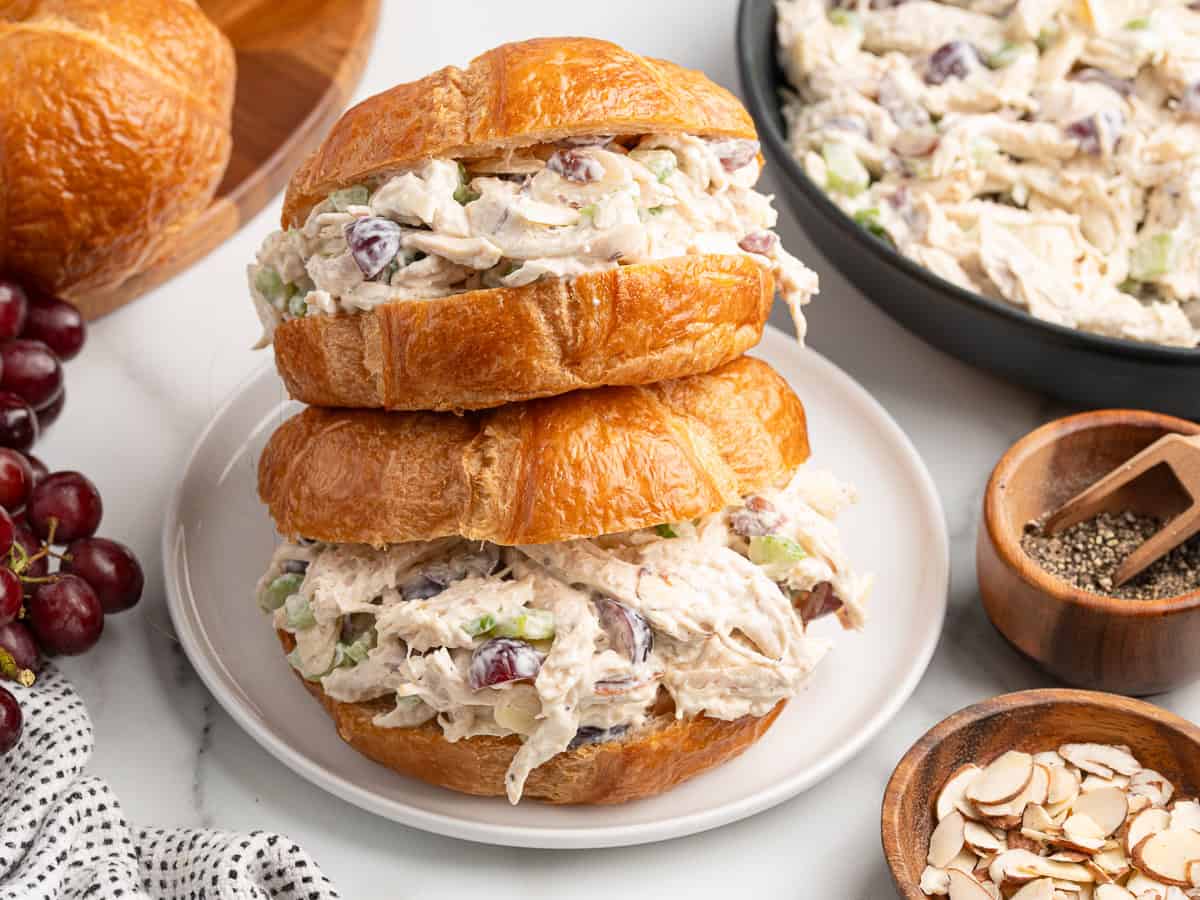 Two Chicken Salad Sandwich croissants filled with creamy chicken salad, grapes, celery, and almonds are stacked on a white plate, surrounded by a bowl of salad, grapes, sliced almonds, and pepper on a marble surface.