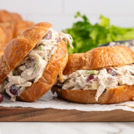 Two croissant Chicken Salad Sandwiches filled with creamy chicken salad, grapes, and celery rest on parchment paper, surrounded by fresh grapes and leafy greens in the background.