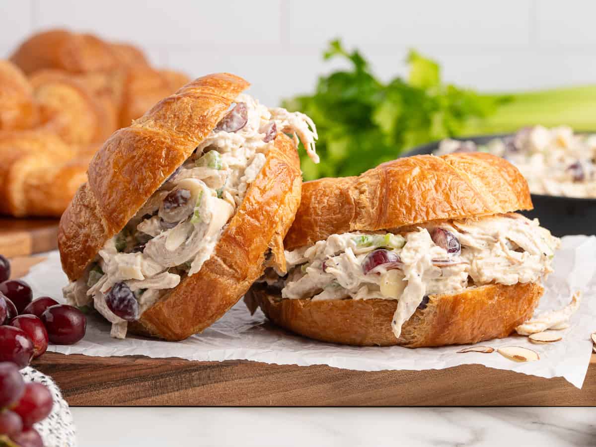 Two croissant Chicken Salad Sandwiches filled with creamy chicken salad, grapes, and celery rest on parchment paper, surrounded by fresh grapes and leafy greens in the background.