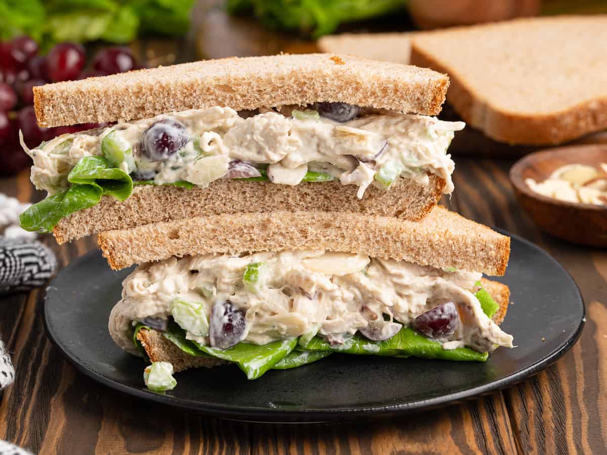 A Chicken Salad Sandwich with lettuce, sliced in half and stacked on a black plate, features visible grapes and almond slices, set on a wooden table with extra grapes, bread, and lettuce in the background.