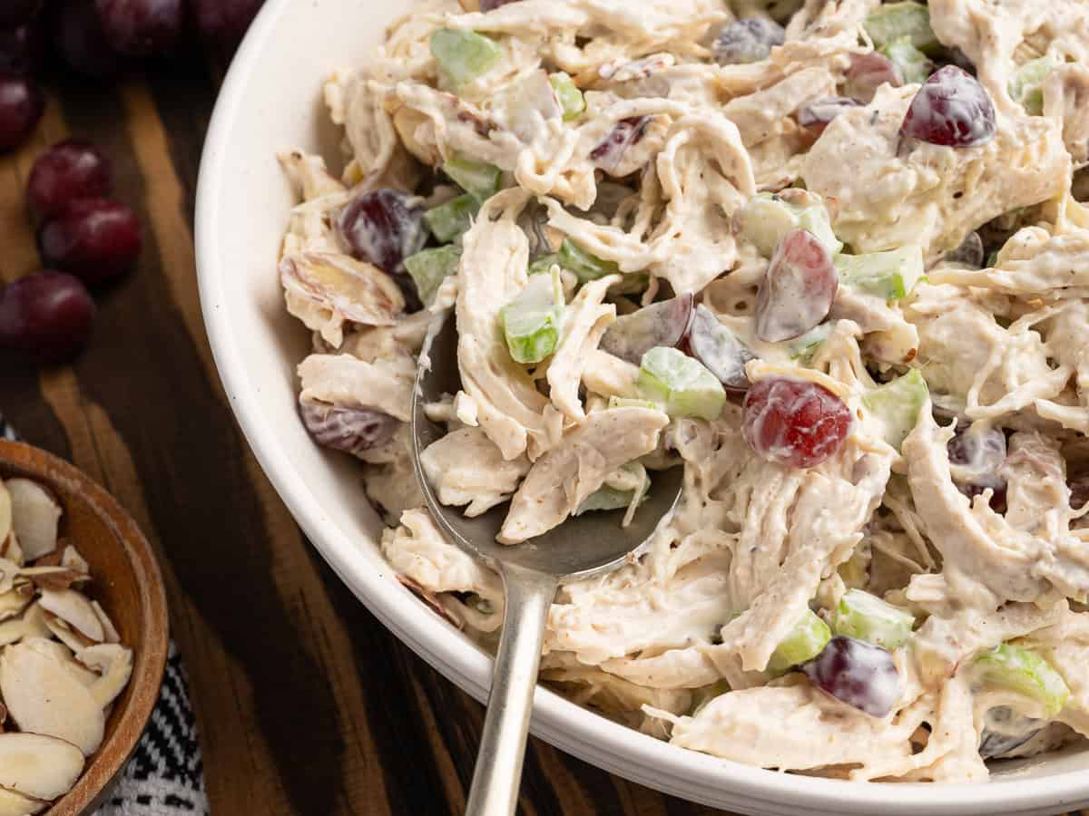 A bowl of chicken salad with shredded chicken, sliced celery, red grapes, and creamy dressing, perfect for enjoying on its own or in a Chicken Salad Sandwich. Sliced almonds and whole grapes are nearby on a wooden table.