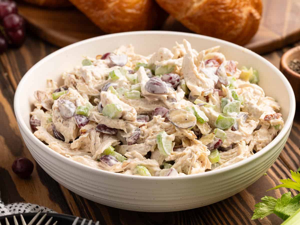 A bowl of chicken salad with shredded chicken, green celery slices, red grapes, and creamy dressing sits on a wooden tableโperfect for spooning onto bread rolls to make a delicious Chicken Salad Sandwich. Bread rolls and pepper are in the background.