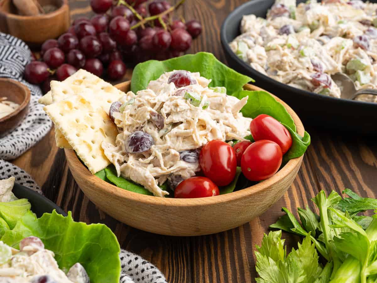 A wooden bowl filled with chicken salad containing grapes, placed on lettuce leaves, with cherry tomatoes and two crackers on the side. A Chicken Salad Sandwich, extra grapes, and a larger bowl of chicken salad are in the background.