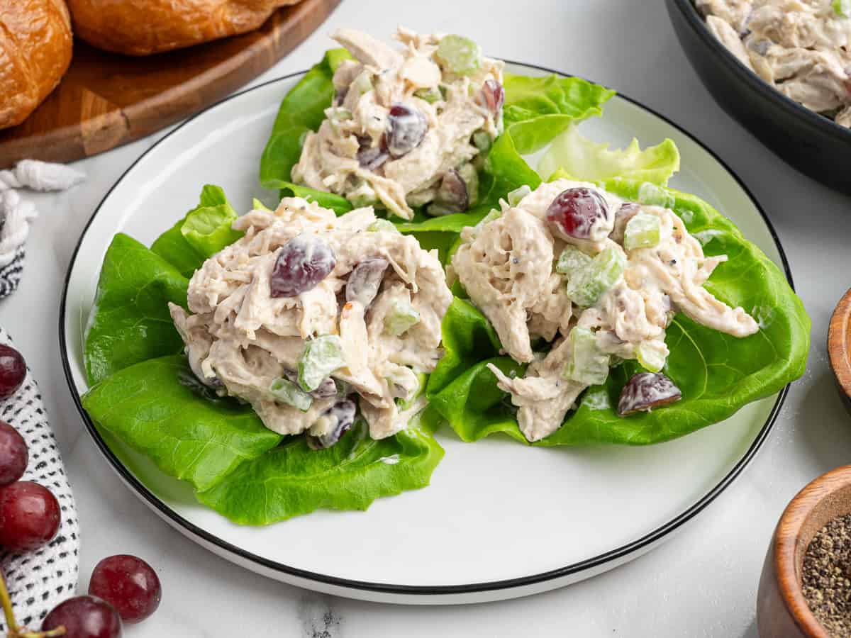 Three lettuce leaves topped with a creamy chicken salad containing grapes and celery are arranged on a white plate, making a light twist on the classic Chicken Salad Sandwich. A bowl of more salad, red grapes, and seasonings are visible nearby.