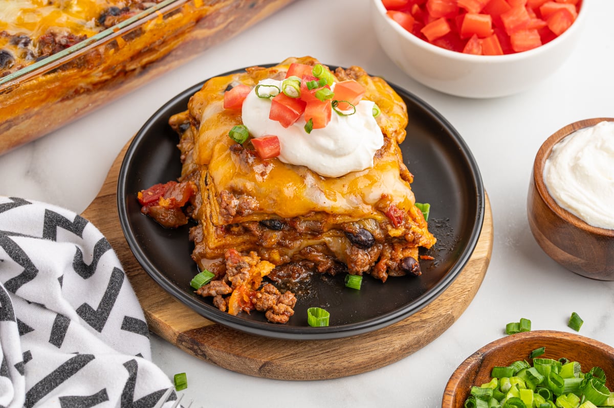 a slice of mexican casserole topped with sour cream and garnished with diced tomatoes and green onions on a plate