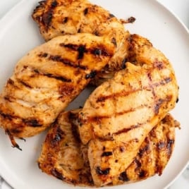 Overhead view of grilled chicken breasts stacked on a white plate, served with forks and a lemon water drink on the side.