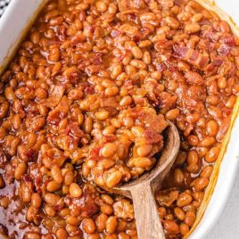 overhead shot of baked beans in a white baking dish being picked up with a wooden spoon