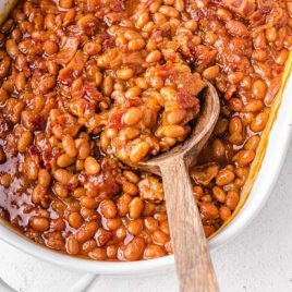 close up overhead shot of baked beans in a white baking dish being picked up with a wooden spoon