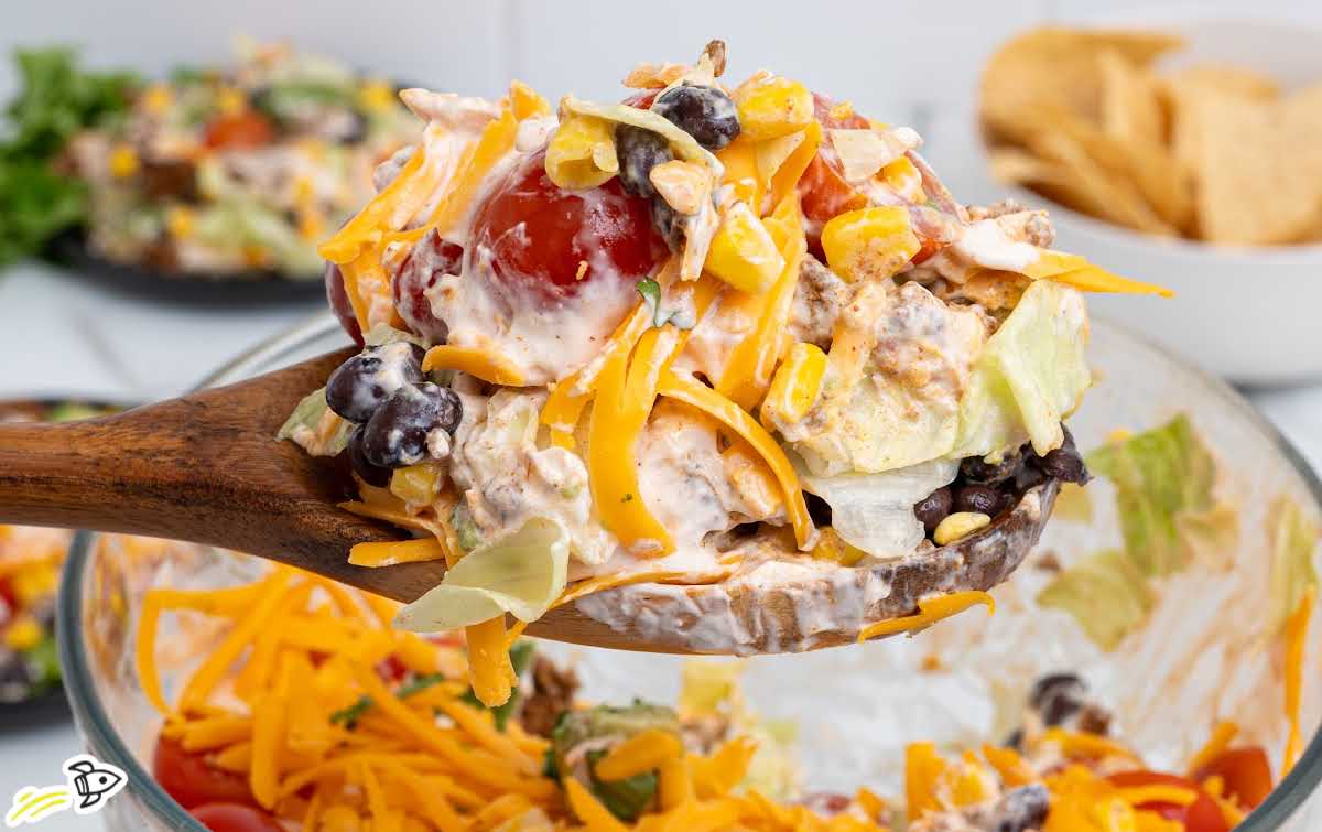 Close-up of layered taco salad served on a wooden spoon with lettuce, beans, corn, beef, tomatoes, cheese, and dressing