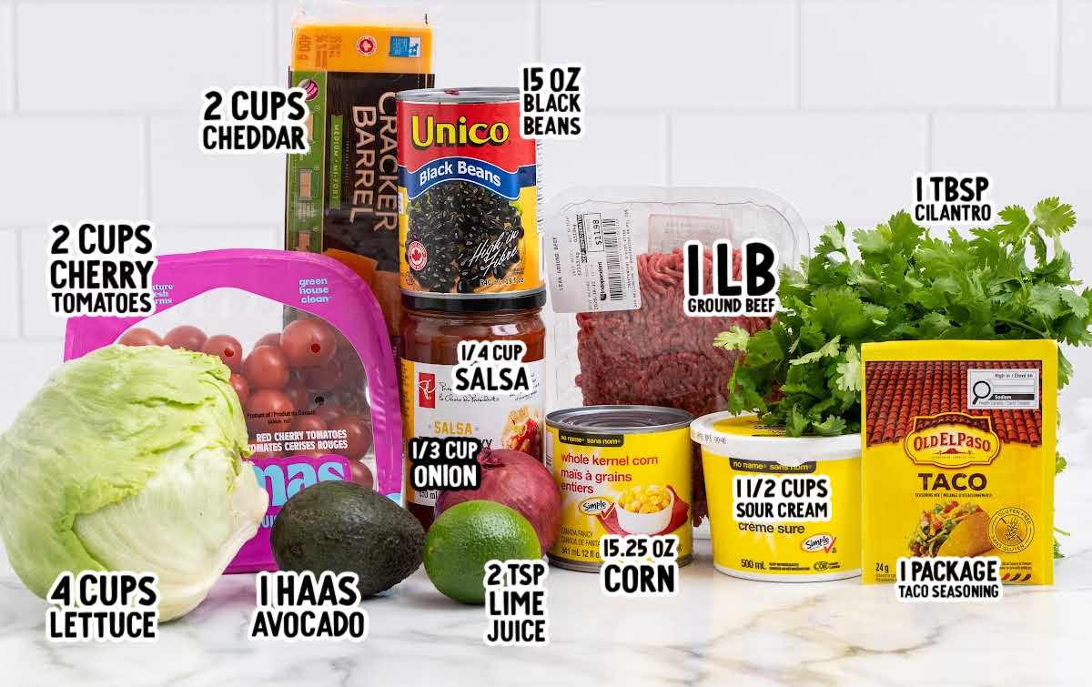 Ingredients for layered taco salad including ground beef, taco seasoning, black beans, corn, cherry tomatoes, lettuce, cheddar cheese, avocado, sour cream, salsa, lime, cilantro, and onion