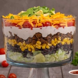 Fully loaded layered taco salad in a clear pedestal bowl with visible layers of lettuce, beans, corn, beef, dressing, tomatoes, cheese, and avocado