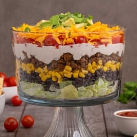 Layered taco salad in glass serving bowl with distinct layers of lettuce, black beans, corn, seasoned beef, creamy dressing, tomatoes, cheese, and avocado