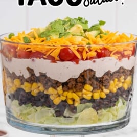Layered taco salad in a glass bowl topped with cheddar cheese, cherry tomatoes, and avocado with text overlay Fully Loaded Taco Party Salad
