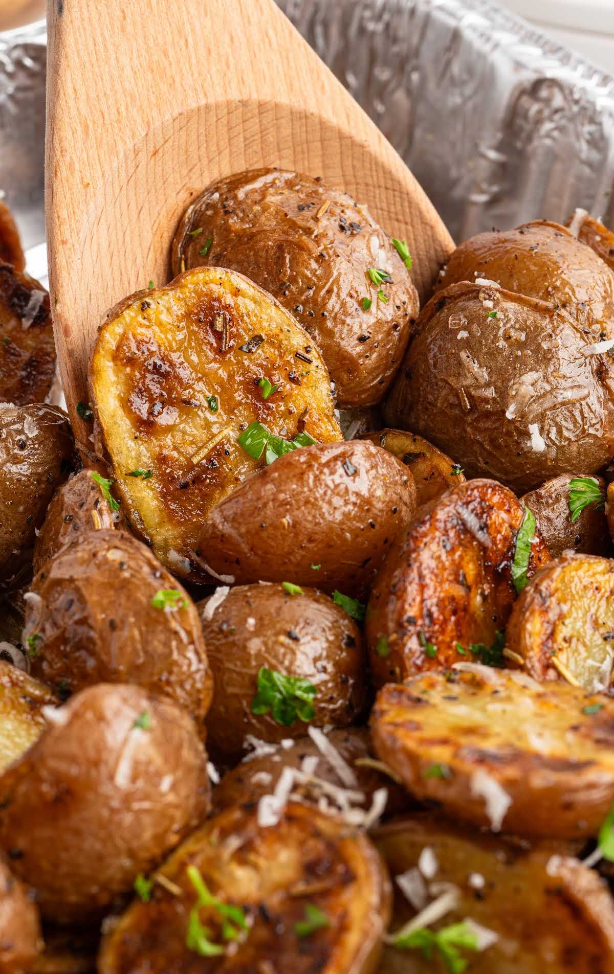 Wooden spoon lifting seasoned grilled potatoes with crispy edges from a foil tray