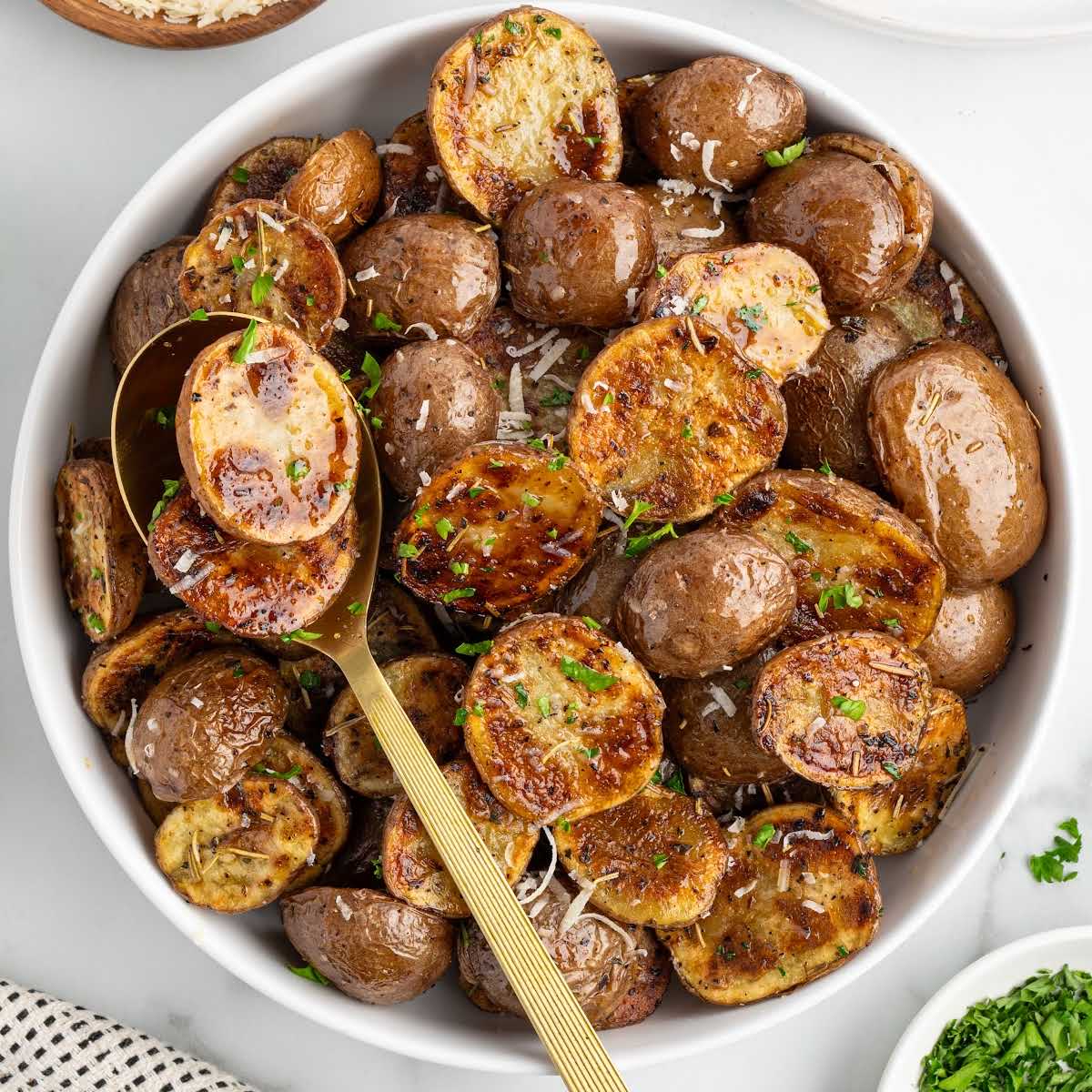 Overhead image of grilled potatoes in a white serving bowl with golden serving spoon and fresh parsley