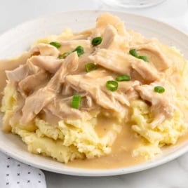 Plate of mashed potatoes topped with shredded crockpot chicken and gravy, garnished with sliced green onions.