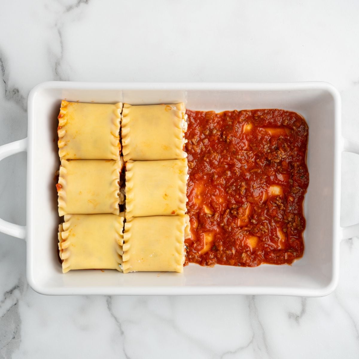 Six Lasagna Roll Ups are placed side by side in a white baking dish, with tomato sauce covering only half of the bottom, leaving the other half beneath the noodles uncovered. The dish is set on a marble surfaceโperfect for your next lasagna roll ups recipe.