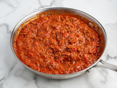 A stainless steel pan filled with thick, chunky red tomato and ground meat sauce for a delicious Lasagna Roll Ups recipe sits on a white marble surface.