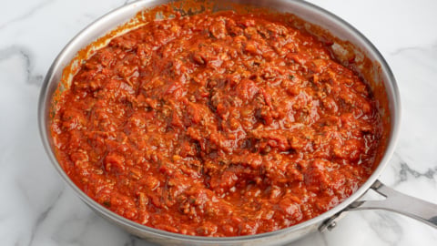 A stainless steel pan filled with thick, chunky red tomato and ground meat sauce for a delicious Lasagna Roll Ups recipe sits on a white marble surface.