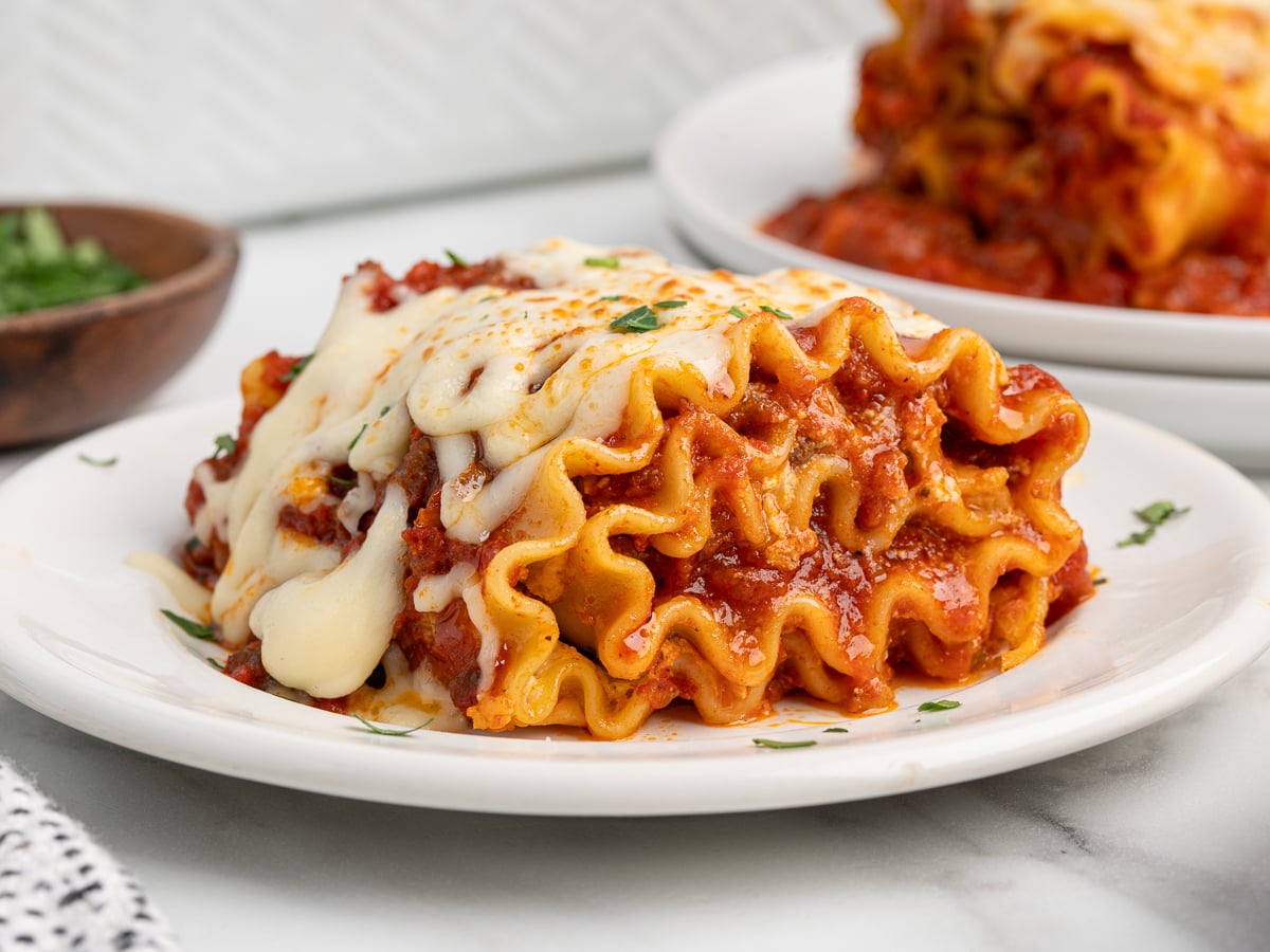 A close-up of a serving of cheesy Lasagna Roll Ups topped with marinara sauce and melted cheese, garnished with herbs, on a white plate. Another delicious lasagna roll ups recipe is visible in the background.