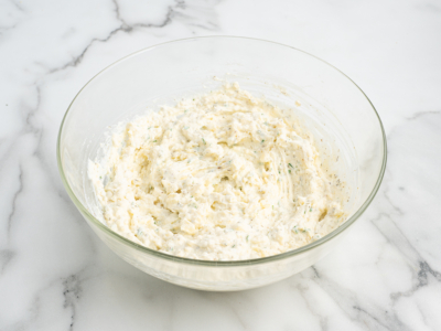 A clear glass bowl filled with a creamy, white cheese mixture with visible herbs, perfect for layering in your favorite lasagna roll ups recipe, sits on a white marble surface.
