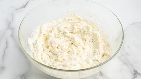 A clear glass bowl filled with a creamy, white cheese mixture with visible herbs, perfect for layering in your favorite lasagna roll ups recipe, sits on a white marble surface.