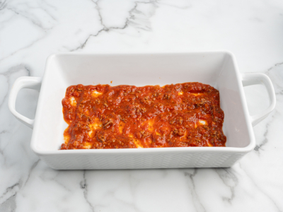 Rectangular white baking dish with handles containing a thin layer of red tomato sauce with visible herbs and small pieces of cheese, perfect for preparing your favorite Lasagna Roll Ups recipe, placed on a white marbled surface.