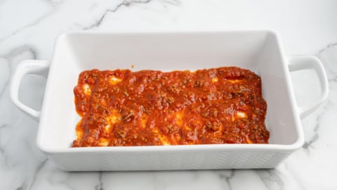 Rectangular white baking dish with handles containing a thin layer of red tomato sauce with visible herbs and small pieces of cheese, perfect for preparing your favorite Lasagna Roll Ups recipe, placed on a white marbled surface.